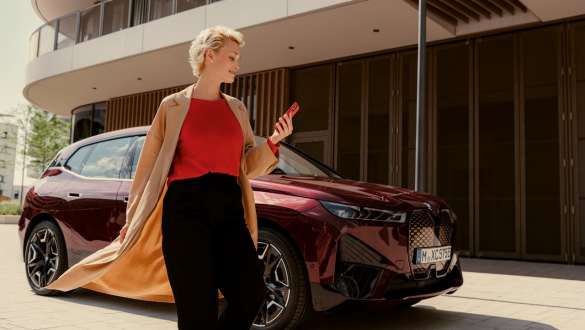 My BMW aplikacija Moja BMw app Žena na ulici sa pametnim telefonom u ruci i BMW vozilom u pozadini