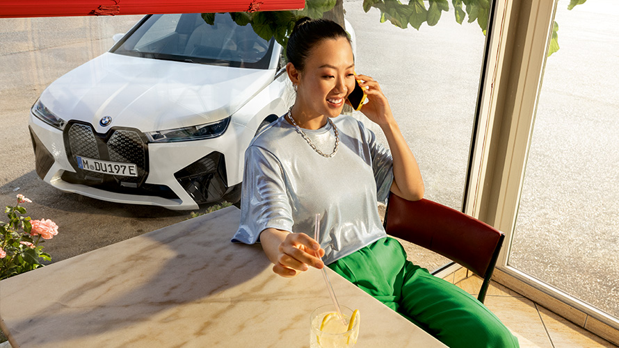 Uslovi Proaktivna nega Proaktivna nega, Provera i održavanje Uslovi Proaktivna nega Proaktivna nega, provera i održavanje Žena sedi za stolom sa pametnim telefonom u ruci i modelom BMW-a u pozadini