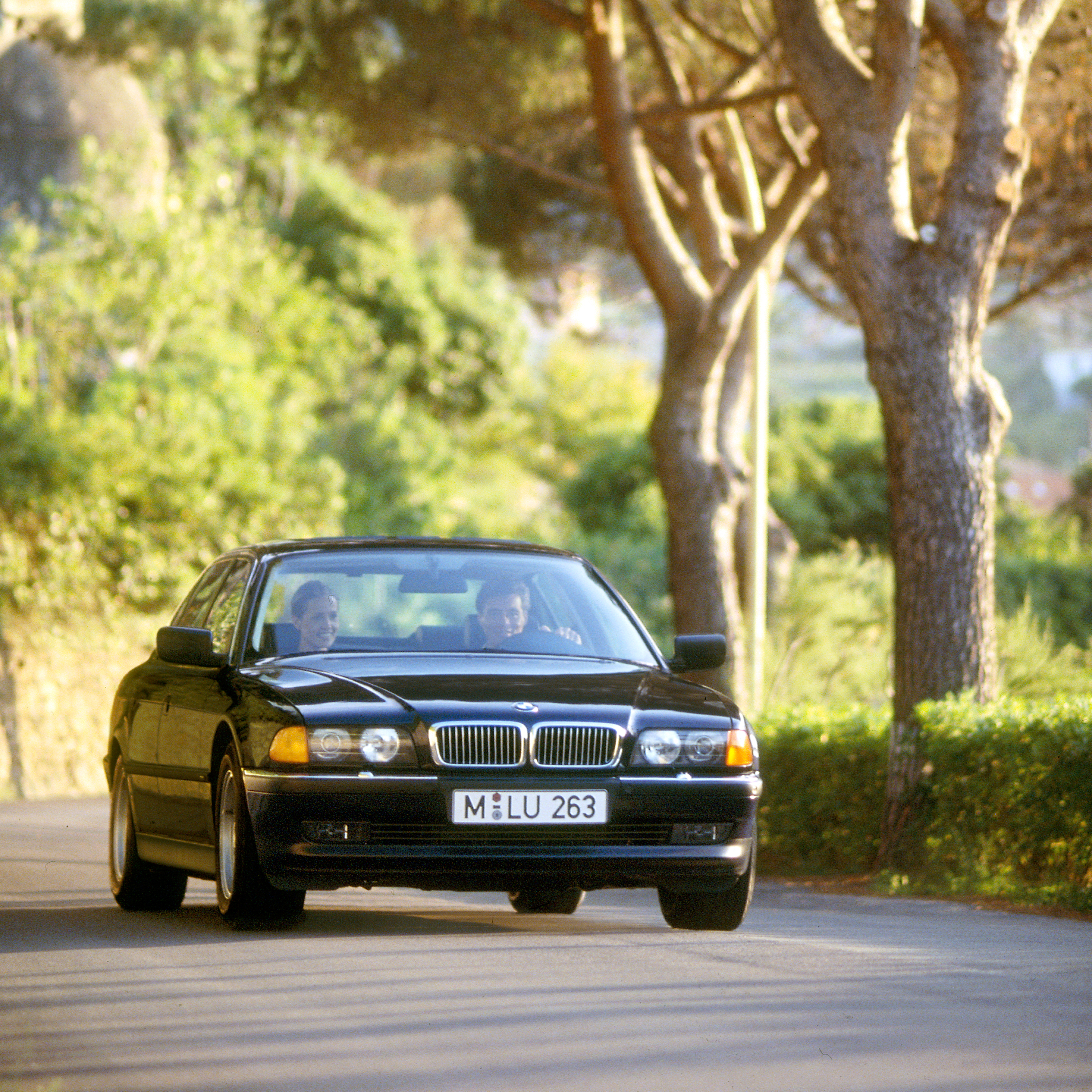 BMW Serije 7 Sedan E38 na aveniji u seoskom okruženju tokom leta sa malim gradom u pozadini